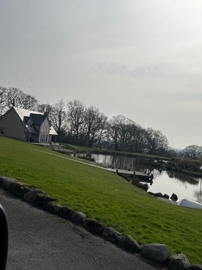 The image presents a tranquil waterside property situated within the Borough of Wyre, United Kingdom, observed during daylight under an overcast sky. In the mid-ground left, a contemporary residence features light-colored siding, a dark slate-grey gabled roof, and multiple windows, including a large bay window facing a body of water. A paved patio with outdoor furniture is positioned directly in front of the house, adjacent to the water's edge. An extensive, well-maintained green lawn slopes from the foreground down towards the house and the water.

A calm, dark-toned body of water, possibly a pond or small lake, occupies the mid-ground, reflecting the sky and surrounding trees. A wooden jetty extends from the grassy bank into the water on the right side of the frame. Further to the right, a small, light-colored boat rests on the grass near the water's edge. The far bank of the water and the background are densely populated with deciduous trees exhibiting bare branches, indicative of a late winter or early spring season. The immediate foreground consists of a dark asphalt surface, presumably a driveway, separated from the lawn by a line of irregular, moss-covered stones. No individuals or animals are present in the scene.