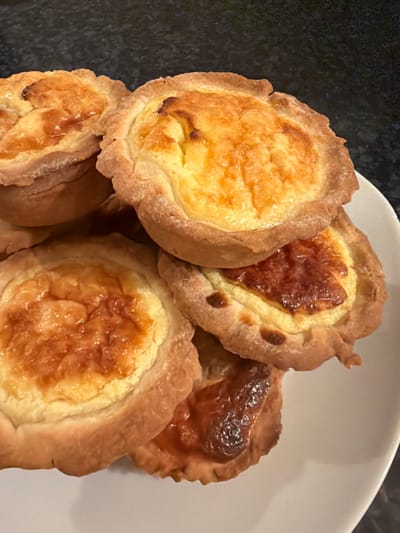 The image displays a close-up, static shot of approximately seven small, circular, baked pastries, resembling tarts or mini quiches, stacked and overlapping on a white plate. Each pastry features a crimped, golden-brown crust, enclosing a creamy, baked filling that ranges in color from light yellow to golden-orange. Several pastries exhibit areas of darker browning or caramelization on their filling surfaces, notably one in the lower right foreground. The white plate, partially visible, rests on an out-of-focus, dark grey or black textured surface, consistent with a kitchen countertop. Illumination appears to be overhead. No individuals, actions, or interactions are depicted. This scene is situated in Givisiez, Switzerland.