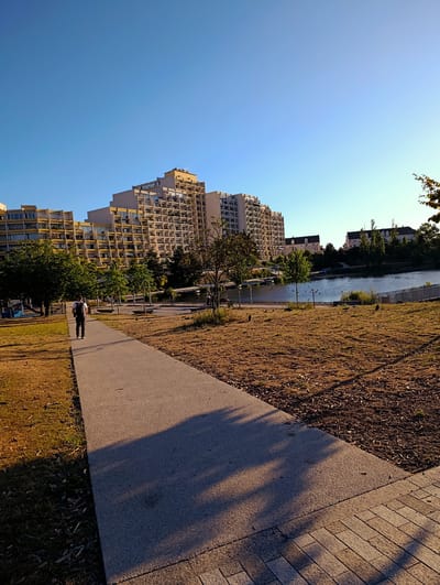 The image depicts a park scene with a large, modern residential building in the background. A paved walkway winds through the park, leading towards the building. A lone individual, a male wearing a backpack, walks away from the viewer on the walkway. The park features sparse, dry grass, scattered trees, and a body of water in the middle ground. Several ducks are visible on the water. The sky is clear and blue, indicating a sunny day. The architectural style of the building suggests it is part of the "Nouveau Monde" residential complex in Noisy-le-Grand, France, known for its distinctive, organic design by architect Ricardo Bofill. The overall impression is one of urban nature and modern living coexisting.