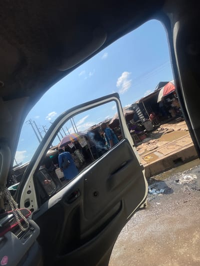 The image depicts an outdoor scene in a market area, likely in Mowe, Nigeria. The viewpoint is from inside a vehicle, looking out through an open car door and the windshield. The foreground is dominated by the interior of the car, with a dark grey dashboard and door panel visible. A beaded garland hangs from the dashboard.
The main subject outside the car is a bustling market. Stalls with colorful awnings and various goods are set up along a wet, muddy ground. Several people are visible, some interacting with vendors, others moving through the market. One person in a blue shirt is seen in the middle of the frame, seemingly engaged in an activity at a stall. The market appears to be in full swing.
The sky is bright blue with scattered white clouds, suggesting it is daytime, likely mid-morning or afternoon. The presence of puddles and wet surfaces indicates recent rain or ongoing water accumulation, typical of market environments after rainfall. Power lines are visible against the sky, hinting at the presence of electricity infrastructure. The overall impression is one of a lively, if somewhat unkempt, local marketplace. There is no visible text in the image.