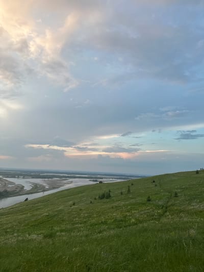 This is a landscape photograph taken from a grassy hilltop overlooking a wide river and its surrounding floodplain. The sky is filled with a mix of clouds, with soft hues of pink, peach, and pale blue suggesting either sunrise or sunset. The foreground is dominated by a lush, green, grassy slope, dotted with a few small evergreen saplings. In the middle ground, the river meanders, with visible sandbars and vegetation along its banks. The background shows a distant, flat horizon with what appears to be a small settlement or trees. On the far right of the hilltop, there are two small, dark, silhouetted figures standing. The overall impression is one of serene natural beauty in the countryside, likely near Nizhniye Vyazovye, Russia, given the context.