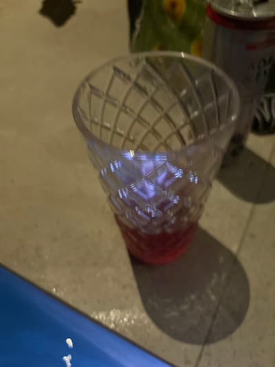 The image shows a clear glass filled with a pink liquid. It is resting on a light beige surface. The glass casts a shadow on the surface. The upper left of the image shows a blue surface, possibly water. The coordinates provided (latitude: 9.647, longitude: -85.166) correspond to a location in the city of Santa Fe, Costa Rica. There is no evidence of any people or other objects in the scene.