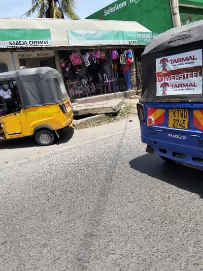 The scene depicts a commercial street in Mkomani ward, Kenya, under clear daylight conditions. Two three-wheeled auto-rickshaws (tuk-tuks) are positioned on the asphalt road. The left vehicle is a yellow auto-rickshaw with a black canopy; its front is partially cropped, revealing a passenger wearing a white top and a head covering seated inside. The right vehicle is the rear section of a blue Piaggio auto-rickshaw with a black canopy, displaying Kenyan license plate "KTWD 274E" and "TARMAL POWERSTEEL" stickers.
In the background, a row of single-story commercial buildings with corrugated metal roofs lines the street, separated by a concrete curb and a narrow dirt strip containing a drainage ditch. Signage identifies businesses including "SABEJA CHEMIST," "MASJID BOMANI," and multiple instances of "Safaricom." An open-fronted shop, displaying various merchandise such as bags, hats, and shoes, is visible between the two auto-rickshaws. Several individuals are present in and around this shop; one person in a red shirt and blue jeans stands near the entrance, and others are seated or standing inside. Palm trees are visible against the blue sky above the buildings. The auto-rickshaws appear stationary.