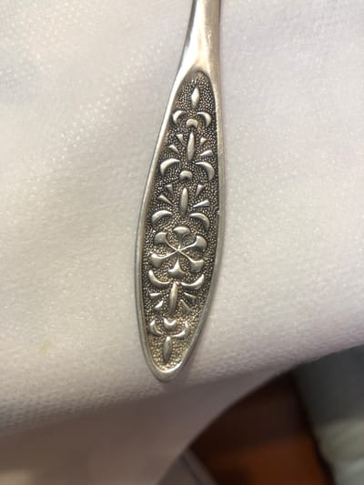 A close-up, overhead shot shows the decorative end of a silver spoon resting on a textured white surface. The spoon's handle is elongated and oval-shaped, featuring an embossed floral pattern. The pattern consists of repeated stylized fleur-de-lis motifs arranged symmetrically along the center of the handle. The background is a soft, textured white material, possibly fabric or paper, with a slight sheen. The lighting creates subtle shadows that highlight the relief of the spoon's design. The location is not discernible from the image.