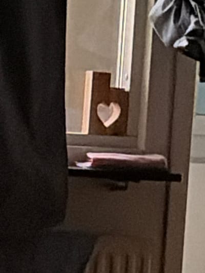 A wooden heart-shaped decoration is placed on a windowsill in Solna, Sweden. The decoration is dark brown and has a cutout heart shape in the center. It is positioned in front of a window with vertical blinds. To the left of the decoration, a black curtain is visible, and to the right, a white or light-colored vertical frame of the window. Beneath the decoration, a thin, dark shelf holds two objects, one appearing to be a stack of papers or a thin book with a reddish cover. The overall scene suggests an indoor setting, possibly a home or office.