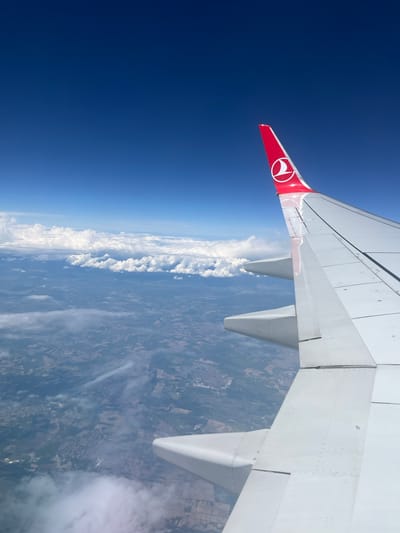 The image shows a view from an airplane window. The wing of a Turkish Airlines plane is visible, with its distinctive red tail fin and white crescent moon and star logo. The sky is clear and blue to the left. Below the plane, the landscape includes fields, towns, and clouds. Based on the landscape, the location of the photo is near the city of Boliartsi, Bulgaria. The terrain exhibits a patchwork of agricultural fields and settlements.