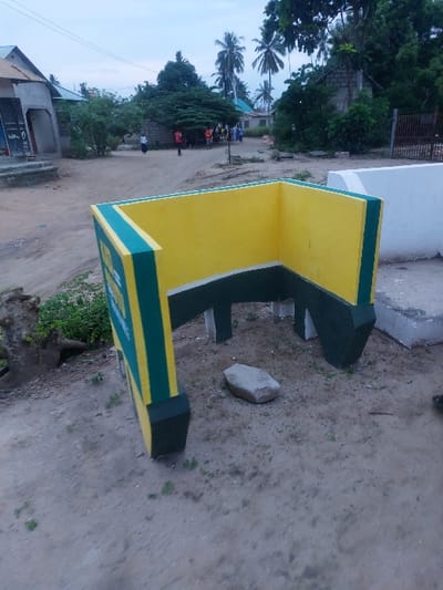 The image displays a street scene in Mwandege, Tanzania, characterized by an unpaved, light brown dirt ground under a pale, overcast sky. In the foreground, a U-shaped concrete structure, open toward the viewer, is prominent. This structure is painted bright yellow on its interior surfaces and dark green on its exterior edges and base. An oblong, light grey rock rests on the dirt ground inside the U-shape. Sparse green vegetation protrudes from the dirt around the structure.
In the midground, a wide dirt path extends into the distance. To the left of the path, a single-story building with a light-colored, sloped roof and walls, featuring a darker entrance, is visible. Further along the path, a group of approximately six to eight individuals is gathered. One person is clearly visible in a red shirt, while others wear varying light and dark clothing. Another individual, dressed in light-colored attire, walks away from this group further down the path.
The background consists of numerous tall palm trees, dense green foliage, and additional residential structures, including one constructed from concrete blocks. A white concrete wall section is visible on the far right. No discernible interactions or specific actions beyond individuals gathering and one person walking are evident.