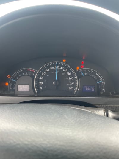This is a close-up, low-angle, vertically oriented shot of a car's dashboard in Kwali, Nigeria. The main subjects are the car's instrument cluster, which includes a speedometer, a tachometer, a fuel gauge, and a digital display. The speedometer is marked in both km/h and mph, with the needle pointing to approximately 110 km/h. The tachometer is marked in rpm, with the needle near the 0 mark. The fuel gauge is showing nearly full. The digital display shows "ODO 89578 km," indicating the odometer reading. There are several warning lights illuminated on the dashboard, including what appears to be an engine light, a battery light, and a brake warning light. The ambient light suggests it is daytime. The overall scene is typical of a car's interior, with the focus on the functional elements of the dashboard.