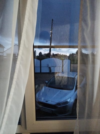 The image captures a view through a window partially obscured by sheer white curtains. Outside, a light-colored car, possibly a Ford Focus, is parked in front of a white, corrugated metal fence with arched gates. The scene appears to be a residential area, with hints of houses and trees in the background. A utility pole with lights is visible in the twilight sky. The overall impression is that of an evening or early morning, indicated by the dim lighting and the dark sky with scattered clouds. The weather seems calm. A license plate on the car is partially visible and appears to read "B636TH36". The setting suggests a suburban or rural location in Sudzhenka, Russia. No people are visible in the frame.