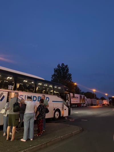 This image depicts an outdoor scene at dusk, likely in Garbsen, Germany, given the context of the prompt. The primary subject is a white tour bus, identified by the large number "181" on its side and the text "SINDBAD" printed prominently. Several people, appearing to be passengers, are gathered near the open door of the bus, some are looking at each other, and others appear to be conversing.
Adjacent to the bus, a line of semi-trailer trucks is parked, some with logos visible, including one that reads "M NACHAN TRANSPORT." Streetlights are illuminated, casting a warm glow on the scene, and the sky is a deep blue, transitioning from twilight. The ground is paved with interlocking bricks and asphalt. The environment suggests a parking area or a rest stop, possibly along a major road or highway. The overall atmosphere is calm, with people preparing to board or disembark the bus.