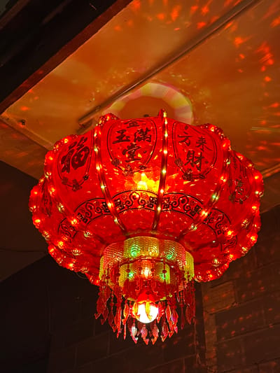 This is a close-up, low-angle shot of a brightly lit, ornate Chinese lantern. The lantern is predominantly red with intricate black patterns and Chinese characters adorning its surface. It is decorated with a string of small, multi-colored LED lights that wrap around its body. At the top of the lantern, there is a section with smaller, decorative elements that appear to be shaped like fish, rendered in red and yellow. The lantern is illuminated from within, casting a warm glow and projecting a pattern of scattered lights onto a textured, yellowish wall in the background. The ceiling above is dark and appears to be made of wood, also showing a scattering of light reflections. The overall impression is festive and possibly celebratory, evoking a sense of tradition and light. The text visible on the lantern includes Chinese characters, likely auspicious phrases or symbols associated with celebration. The scene is dimly lit, suggesting it might be nighttime or an indoor setting with artificial lighting.