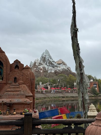 The image depicts an outdoor scene in the city of Bay Lake, Florida, specifically within Disney's Animal Kingdom theme park, focused on the Asia section featuring Expedition Everest. An overcast sky dominates the upper portion of the frame. In the left foreground stands a large, multi-tiered reddish-brown stone structure resembling a shrine, featuring intricate carvings, arched niches (one containing a blue and gold figure), and a flat surface displaying offerings including small fruits, a brass vessel, and a red pot. A dark wooden railing with decorative cutouts crosses the bottom foreground. To the right, a tall, weathered wooden pole is draped with numerous tattered, faded gray fabric strips.

A calm body of water spans the midground, reflecting the sky and distant elements. Three small, brightly colored boats (blue, red, yellow/green) are partially visible at the water's edge, seemingly overturned or resting. Beyond the water, a terraced area with stone steps leads to an outdoor dining or rest area. Multiple individuals are present, seated at tables under numerous red patio umbrellas. Colorful prayer flags are strung between posts in this area. In the far background, a large, artificial snow-capped mountain range, identifiable as Expedition Everest, rises prominently against the gray sky, flanked by trees at its base.