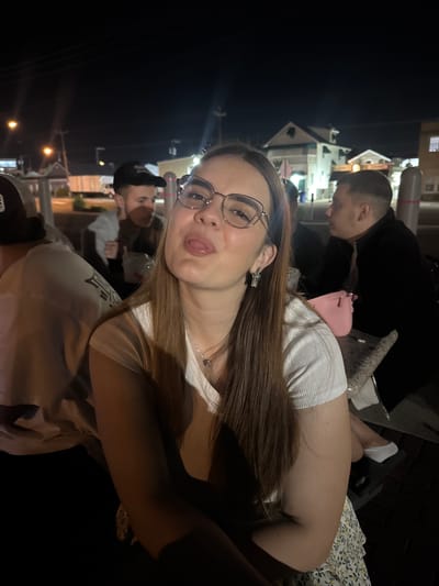 A woman in a white shirt and a floral skirt is sticking her tongue out at the camera while wearing glasses. She has long brown hair and is wearing a heart-shaped necklace and a butterfly earring. A pink purse is visible on a table next to her. In the background, there are other people seated at tables and a street scene with buildings and lights at night. The image is taken in Ocean City, United States of America.