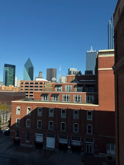 A cityscape under a clear blue sky. Prominent skyscrapers include a green-tinted building, a pyramid-shaped structure, and a slender spire. A brick building with repeating window patterns dominates the foreground. The building casts a shadow on its side. The image has been rotated so that the sky and buildings appear vertically oriented.