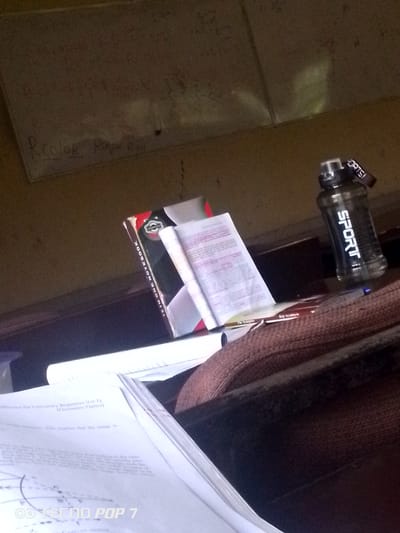 In Eleweran, Nigeria, a cluttered desk is shown, bathed in soft light. A "12-in-one Notebook" lies open, its pages filled with text and diagrams. Next to it, a black water bottle with "SPORT" in white letters stands upright. The desk surface is dark and worn, with papers scattered about, suggesting a study or work area. A brown fabric object, possibly a bag or a cushion, is partially visible in the foreground, adding to the sense of disarray. The background features a faded whiteboard with faint scribbles. The overall impression is one of a busy, but somewhat unorganized, academic or creative space.