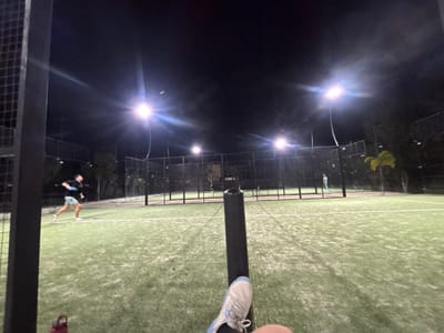 The scene depicts a nighttime view of an illuminated padel or tennis court in Komchén, Mexico. The perspective is low, from the ground level of the court. In the immediate foreground, the right foot and lower leg of an observer are visible, wearing a light grey and blue athletic shoe. A black vertical support post, possibly part of the court's net or a perimeter structure, extends upwards from the bottom center, partially obscuring the view. To the lower left, a maroon water bottle rests on the green artificial turf.
On the court itself, two male individuals are actively engaged in play. A player on the left side, wearing a dark shirt and light-colored shorts, is captured in motion, mid-swing with a racket. A small, light-colored object, consistent with a padel or tennis ball, is suspended in the air near him. Further back, closer to the net, a second player in a lighter-colored outfit is visible. In the far background, a third faint figure is discernible on an adjacent court.
The playing area features green artificial turf with white boundary lines. The court is enclosed by black metal mesh fences and transparent glass panels. The scene is brightly lit by multiple artificial floodlights mounted on tall poles, casting prominent lens flares across the dark night sky. Trees and foliage, including some with distinct yellow-green leaves, are visible beyond the court enclosures in the background.