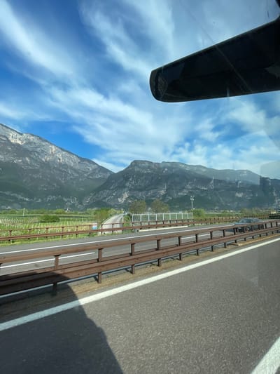 The image, taken from inside a vehicle, shows a view of Zambana, Italy, through the windshield. A section of the vehicle's rearview mirror is visible on the left. Outside, a mountain range dominates the landscape. To the right, a multi-lane highway runs alongside a green area, possibly vineyards, separated by fencing and barriers. The sky is blue with scattered clouds.