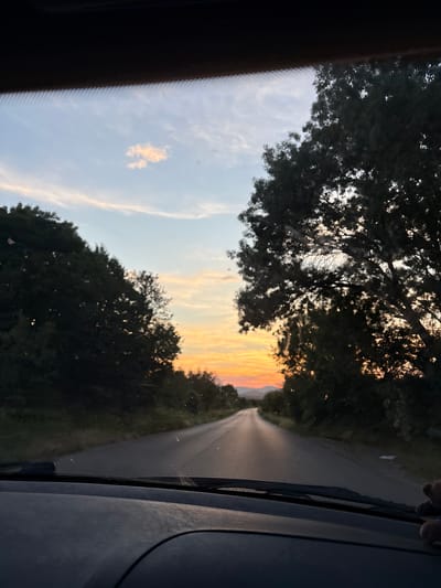 A road curves through a rural landscape at sunset, viewed from inside a vehicle. The sky displays gradients of blue, orange, and yellow, with a few wispy clouds. Trees line both sides of the road, casting shadows and silhouetting against the colorful sky. Distant mountains are visible on the horizon. The foreground shows a portion of the car's dashboard and windshield wipers, suggesting the perspective of a driver. No people or specific structures are clearly identifiable within the scene.
