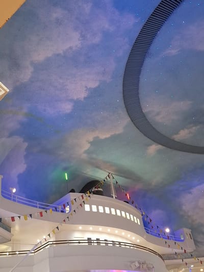 The image shows a portion of an indoor decorative structure designed to resemble a cruise ship, situated in the city of Trafford, United Kingdom. The ceiling above the ship structure is painted with a sky mural, complete with clouds and small, scattered lights that simulate stars. A large, circular architectural element, possibly a ventilation or lighting fixture, is visible against the painted sky. The ship itself is predominantly white, with multiple decks and windows. Small, colorful triangular flags are strung along the railings of the ship's decks, adding a festive touch. Lighting is present on the ship, including spotlights and illuminated windows. The overall impression is of a theatrical or themed indoor environment.