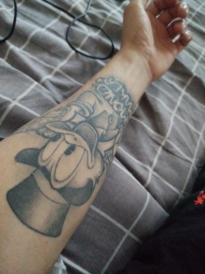 A close-up shot focuses on a person's forearm, which features a tattoo of a Disney character, Goofy. The tattoo depicts Goofy wearing a top hat, seemingly rendered in a grayscale palette. The tattoo extends from the lower part of the forearm towards the wrist, where there is a band of text. The forearm rests on a surface covered with gray and white checkered fabric, possibly a bedspread or blanket. A black cable is also partially visible in the upper left portion of the frame, while a black garment is visible in the bottom right corner of the frame. The location is unspecified, but it is unlikely the image provides any information about being in Gus-Khrustalny, Russia.
