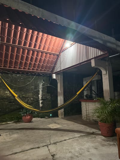 The image displays a dimly lit outdoor or semi-enclosed patio area at night in Tres Valles, Mexico. A yellow and black striped hammock is suspended low across the center, attached to an aged, textured concrete wall on the left and a concrete support pillar on the right. The floor is a concrete slab with visible cracks and some light organic growth. Two potted plants are present: a smaller green plant in a terracotta pot is positioned on the left, near the wall, and a larger, leafy green plant in a similar pot is on the right, beside another concrete pillar.

Above, a red corrugated metal roof, designed to mimic roof tiles, covers a portion of the area. An open section in the overhead structure reveals the dark night sky, with a corrugated galvanized metal wall section visible through the opening. Concrete beams and columns form the structural support. Wires are horizontally strung below the red roof and across the left wall. A cylindrical structure with a red top rim, possibly a well or raised planter, is partially visible on the far right. No individuals are present in the scene.