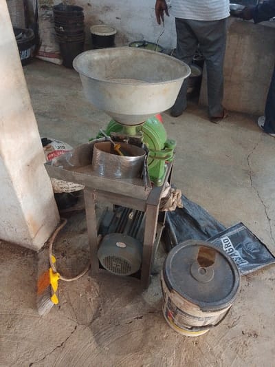 This image depicts an indoor scene in Olomoda, Nigeria, featuring a grain or feed grinding machine as the central subject. The machine comprises a large, silver-colored conical hopper atop a green-painted main body, which connects to a metal framework supporting an electric motor at its base. A perforated metal sieve component is visible on a flat tray section of the machine, with a yellow-handled tool resting inside it.
Two individuals are partially visible in the background, primarily from the waist down. One person, on the left, wears dark trousers and sandals, with their right arm and hand extended over the grinding machine. Another person, on the right, wears dark trousers and white shoes. They are standing behind the machine, observing the process or awaiting operation.
The environment consists of a light-colored concrete floor with visible cracks and stains. A concrete pillar or support structure is positioned on the left side of the frame. In the background, against a light-colored wall, multiple dark-colored buckets are stacked, and a white bucket with a lid is also present. Adjacent to the grinding machine, a large white bag with red lettering, indicating "50kg" and "LAYER FEED 20%", is partially visible. On the floor to the right, a black woven sack labeled "FISAGRO" is spread out. Further right, a white bucket with a multi-colored label, topped with a dark lid featuring a large irregular opening, stands on the floor. A broom with a yellow handle and brown bristles leans against the concrete pillar on the left, connected by a beige electrical cord that runs towards the machine. The scene suggests an active processing or manufacturing area.