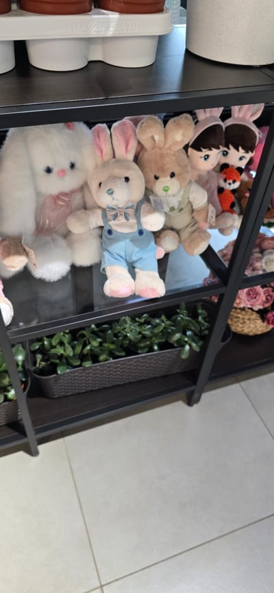 In a retail setting, possibly a shop in Русская Деревня-2 Кп, Russia, a black metal shelving unit holds various items. The top shelf displays white planters and a large white stuffed bunny with a pink ribbon. Below that, a second shelf is filled with several stuffed animals, including a white bunny in blue overalls and a bow tie, a brown bunny in a vest and shirt, and other assorted plush toys. The bottom shelf contains a dark brown woven planter filled with lush green plants. A woven basket filled with pink roses is visible on the right side of the bottom shelf. The overall environment appears to be well-lit, suggesting an indoor retail space.