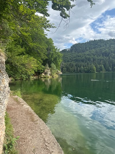 The image shows a serene lake scene, likely in the vicinity of Kufstein, Austria, given the lush forested mountains that surround the water. The foreground features a weathered stone embankment and a gravel path leading to the clear, green water. The lake is calm, with reflections of the sky and surrounding trees shimmering on its surface. In the distance, a densely wooded mountain slope rises majestically, its peak partly obscured by dramatic, cloud-filled skies. A lone individual is visible in the lake, swimming towards the right of the frame. The overall impression is one of tranquility and natural beauty.