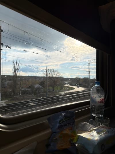 The image, apparently taken from inside a moving train, shows an outside view through a window. Beside the window on the right are a bottle of "Svyatoy Istok" water, and a bag of Dr. Korner chips. The view outside is of a rural landscape including railway tracks, transmission towers, trees, buildings, and roads. The sky is partly cloudy. The train is traveling through or near the city of Krasnyy Sulin, Russia, as evidenced by the brand of chips.