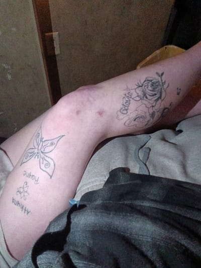 This scene is situated in Ozark, United States. The image depicts a close-up, downward-angled view of a person's bent right leg, from the mid-thigh to the upper calf. The skin is fair-toned.

On the outer right thigh, above the kneecap, an outline tattoo features two roses with leaves, accompanied by the vertically oriented name "Octavia." A small, dark, undefined mark is visible further up the outer thigh. On the inner/front aspect of the upper calf, below the kneecap, a large outline tattoo of a stylized butterfly design is present. Below this, arranged vertically, are the outline names "Rayne" and "Athyna," with the latter positioned next to a small, outline floral or star-like motif.

The kneecap and surrounding skin exhibit several reddish-brown, circular marks and a localized purplish discoloration, consistent with minor skin trauma or bruising. The leg is partially obscured by two layers of clothing: an upper layer of light grey fabric, consistent with sweatpant material, on the upper thigh, and a lower layer of dark grey or black fabric, resembling denim, covering the lower leg.

The background is out of focus, revealing a muted brown wall surface. A vertical wooden trim and a white rectangular object, likely a light switch or outlet, are visible on the left side of the background. A faint yellowish object is partially discernible in the upper right background.