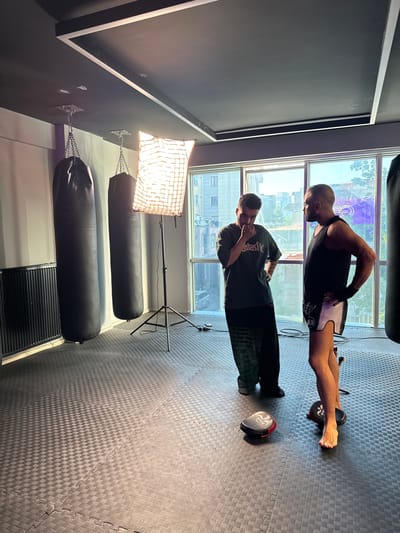 Two men are in a gym with padded flooring and large black punching bags hanging from the ceiling. The scene is illuminated by a large softbox light. One man, wearing a black tank top and white shorts with black trim, is doing a plank exercise with his feet resting on focus mitts. The other man, wearing a dark t-shirt with "Gilas.lk" visible in white text, stands facing the first man with his hands up as if to spar. The gym has large windows that look out onto an urban environment, suggesting it is daytime. The overall atmosphere appears to be one of training or preparation.