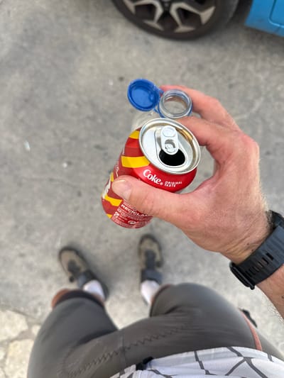 The image shows a person's hand holding an opened can of Coca-Cola and a plastic bottle with its cap removed, seen from a first-person perspective. The background is a paved surface, likely a street or sidewalk, with faint shadows. The person is wearing athletic shorts and socks, and a smartwatch is visible on their wrist. The environment appears to be outdoors in a city setting, given the presence of asphalt and the suggestion of urban elements like a vehicle wheel and car door in the background. The specific location is identified as La Llacuna, Spain.