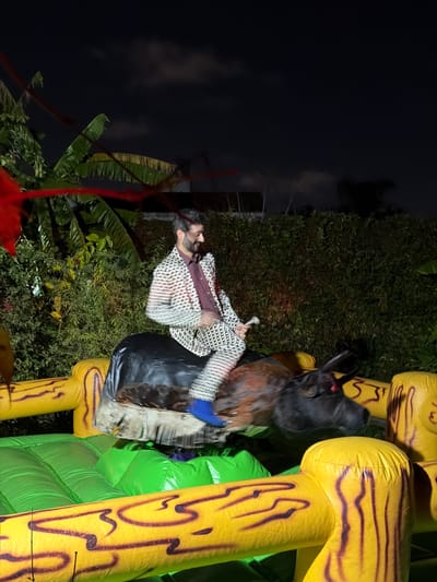 A man is on a mechanical bull that's positioned on an inflatable bounce house outdoors at night. He's wearing a patterned jacket with a maroon collar. The bull is black and brown with red eyes and protruding horns. The bounce house is green and yellow. Green plants and foliage surround the scene. Dark sky is visible in the upper left corner. The location is Florida, Argentina.