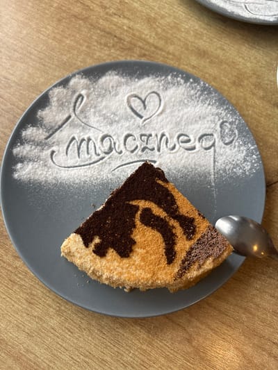 A slice of tiramisu sits on a gray plate dusted with powdered sugar, forming the word "macznego" and a heart. A spoon rests on the right side of the plate. The scene is set on a wooden table. The image was taken in Hajnówka, Poland.
