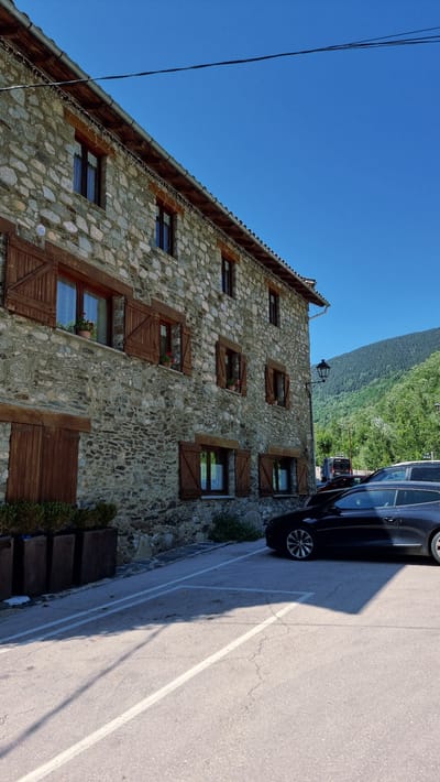 The image captures a stone building with wooden shutters and windows in Setcases, Spain. The building appears to be a traditional house or guesthouse, with several floors and visible stonework. In front of the building, there is a parking area with several cars, including a black hatchback parked prominently in the foreground. The environment is characterized by a clear blue sky and a lush green hillside in the background, indicating a rural or mountainous setting. The sun is shining, casting shadows on the ground. There are no people visible in the image.