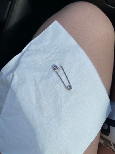 A safety pin rests on a white paper napkin, which is placed on a person's leg. The background is dark, suggesting an indoor or shaded environment. There are no specific details to indicate the location in the city of Synhai, Ukraine.