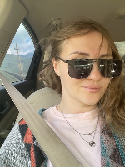 In a car, a woman with blonde hair and sunglasses is looking at the camera. She is wearing a pink shirt and a scarf. The car is moving through a mountainous area with a cloudy sky.
