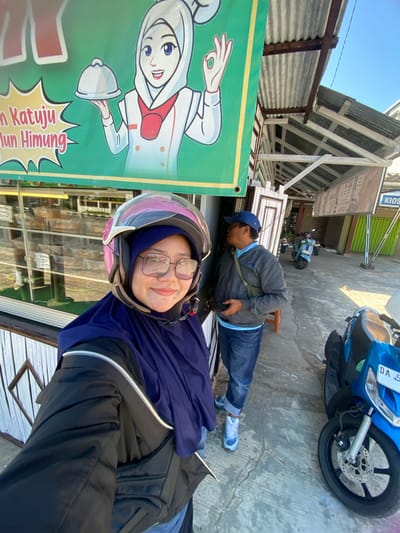 In Sungai Paring, Indonesia, a woman wearing a pink helmet and a navy blue hijab poses for a selfie in front of a storefront. She is smiling and wearing glasses, a black jacket, and a navy blue outer garment. Behind her, a man in a blue baseball cap and grey jacket looks away from the camera, holding a black smartphone. A sign featuring a cartoon chef with a hijab hangs on the wall behind them. Scooters are parked in the background. The environment appears to be outdoors, possibly a street or market area, with a concrete ground and corrugated metal roofing visible.