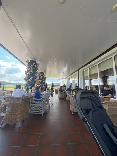 The image depicts an outdoor seating area of a cafe, possibly in Akhlebaikha (Kovriginskii S/S), Russia, given the context. The area is covered by a light-colored roof, supported by columns. A floor made of reddish-brown square tiles is visible.

In the foreground, a dark grey baby stroller is positioned. To the left of the stroller, a man in a blue shirt is seated at a table, looking down at his phone. Further left, a man in a white shirt is also seated at a table.

Several tables with woven wicker chairs are arranged throughout the seating area. Some tables are occupied by people dining or conversing. A large decorative column adorned with colorful flowers, sunflowers, and baskets is a prominent feature in the mid-ground.

Beyond the covered seating area, a glimpse of an outdoor landscape with greenery and a cloudy sky is visible. Large windows line the back of the seating area, reflecting the sky and surroundings. Several individuals are seen standing or walking in the background, including a person in a dark uniform and a young child. The overall scene suggests a relaxed, casual atmosphere at an establishment with outdoor dining.