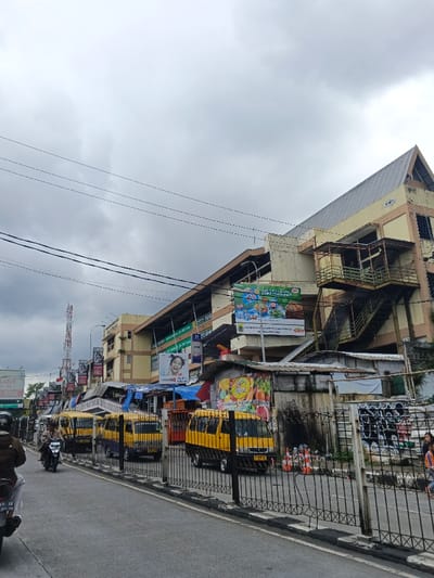 The image depicts an urban street scene in front of the Palayan City Public Market Complex in Palayan, Philippines, under an overcast sky. A paved asphalt road occupies the lower left and central portions of the frame, bordered on the right by a low black metal fence. Several vehicles are present: a person wearing a brown jacket and helmet rides a motorcycle away from the viewer on the left side of the road, followed by another motorcycle. To the right, along the curb, a line of yellow passenger vans with blue trim, resembling mini-jeepneys, are either parked or slowly moving. Orange traffic cones are visible on the ground behind the fence.
Dominating the right side of the image is a multi-story commercial building complex. The building on the far right features a prominent gray gable roof, light yellow walls, and several external balconies with metal railings, connected by an external metal staircase. Further left, additional sections of the complex display multiple large billboards and signs, including advertisements for consumer goods, food products, and one featuring a smiling child. Below these structures, at street level, are smaller, makeshift market stalls constructed from corrugated metal and tarpaulins, one of which displays a large banner with images of fruits and vegetables.
Numerous black electrical wires traverse the upper half of the image against the cloudy sky. In the mid-distance on the left, a red and white striped telecommunications tower is visible above the buildings. A few indistinct human figures are discernible on the far right near the fence and market stalls.