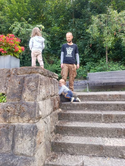In this medium shot, a father stands near a stone structure, observing his two young children. A boy, crawling on stone stairs in the foreground, looks up towards the camera with a curious expression. To the left, on top of the stone structure, a young girl stands with her back to the camera, looking into the distance. The father, with a shaved head and a beard, is dressed in a black t-shirt with the text "DIE IN BATTLE AND GO TO VALHALLA" and light brown cargo pants. The scene is set outdoors, with lush green trees and foliage forming the backdrop. Red flowers in a planter are visible on the left. A wooden bench is situated in the background on the right. The lighting suggests it is daytime, possibly late afternoon given the soft light. The overall atmosphere appears relaxed and familial. The location is identified as Luhačovice, Czechia.