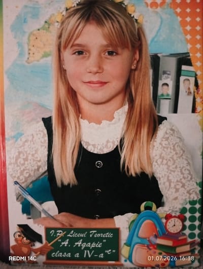 This is a photograph of a young girl with blonde hair, wearing a white lace top with a black dress. She has a flower crown on her head. She is smiling at the camera. In front of her is a small blackboard with text written on it. Behind her, there is a world map and what appears to be a bulletin board with pictures of children. To her right, there is a stack of books with a red alarm clock on top, and a backpack. The bottom of the image contains the text "REDMI 14C" and a date and time stamp "01.07.2026 16:38". The text on the blackboard reads "7.Pl. Liceul Teoretic "A. Agapie" clasa a IV-a"e"". The image appears to be a school photograph. The setting is indoors, likely a classroom or school photography studio. The girl looks happy and is posing for the picture.