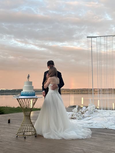 In Osipovo, Russia, a newlywed couple embraces beside a wedding cake on a table. The bride, in a white strapless gown with intricate detailing, holds the groom, who is dressed in a black suit. The three-tiered cake is adorned with a blue gradient and a cake topper of a couple dancing. In the background, a faint rainbow is visible in the pastel-colored sky. The scene unfolds on a wooden deck overlooking a body of water, with a backdrop of trees and buildings. A decorative floral arrangement and a sheer beaded curtain complete the wedding setup.