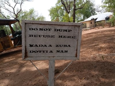 A sign in Rumnde Waziri, Nigeria, at coordinates 9.185, 12.523, reads "DO NOT DUMP REFUSE HERE KADA A ZUBA DOTTIA NAN". The sign is made of metal and is mounted on a metal pole. The sign is in a dirt lot, and a small yellow vehicle is parked nearby. Behind the sign is a building with a tin roof. The building has a window and door. There are trees on the left side of the image. The ground is covered in dirt and grass. The sun is shining. The sign is partially obscured by the vehicle. The sign is positioned so that it is facing the road.