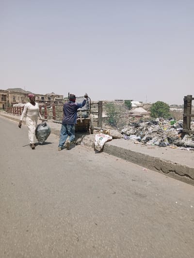 The image captures an outdoor urban scene in Mumbwa, Zambia, under a bright, hazy sky. An asphalt road occupies the foreground and left side. Two individuals are present on or immediately adjacent to this road. On the left, a woman with dark skin wears a light-colored long-sleeved outfit and a dark red head covering. She walks from left to right, carrying a large, dark-colored trash bag in her left hand. To her right, positioned slightly behind her, a man with dark skin wears a dark blue long-sleeved shirt, blue jeans, and a dark red head covering. He stands next to a light-colored utility vehicle or cart with a visible metal frame, his right arm raised towards the frame. Adjacent to the road's edge and behind a concrete curb, a significant accumulation of refuse, consisting of various materials, is piled. A white sack with red lettering rests on the curb near the trash. In the mid-ground and background, multiple light-colored buildings, some appearing dilapidated, are visible, interspersed with sparse green vegetation. A metal railing structure is present behind the man, suggesting the scene takes place on a bridge or elevated roadway.