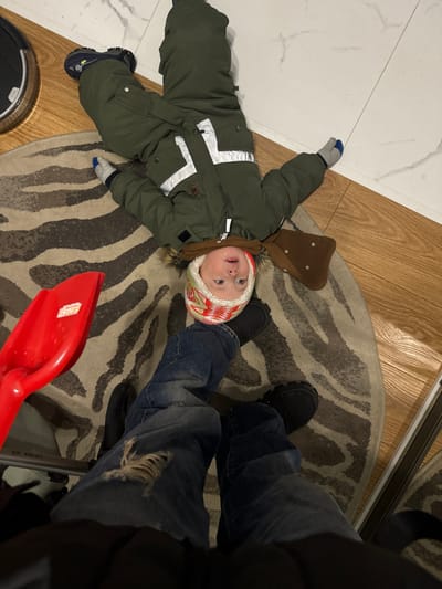 A young child in a green snowsuit and colorful hat lies on a zebra-striped rug. A brown floppy hat is near the child's head. Adjacent to the rug are a hardwood floor and white tiled floor. Nearby is a red toy shovel. A person wearing blue jeans and black shoes stands partly on the rug, partially obscuring the scene. A robotic vacuum cleaner is visible in the corner.