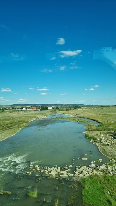 This is a landscape photograph taken from an elevated vantage point, looking down at a river flowing through a rural area. The sky is a vibrant blue with scattered white clouds. In the distance, a small town with red-roofed buildings is visible nestled amongst rolling green hills. The river itself is wide and somewhat shallow, with a rocky bottom visible in places. The banks of the river are lined with green grass and vegetation, with some areas of rocky terrain. A small cluster of white stones are arranged in the river, creating a slight ripple effect. The overall impression is one of a peaceful, rural setting on a clear, sunny day. The location is identified as Milișăuți, Romania.