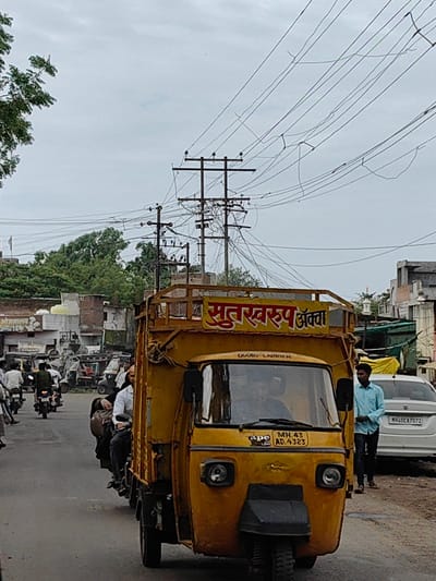 This is a daylight, eye-level, full shot of a busy street in Malkapur, India. In the foreground is a yellow auto-rickshaw, also known as a three-wheeler, with a cargo extension. The auto-rickshaw has a yellow banner with red lettering on top. The license plate reads "MH 43 AD.4323". A man in a light blue shirt and dark pants stands to the right of the auto-rickshaw, looking towards the left. Behind the auto-rickshaw, a white car is parked on the right side of the street. In the midground, several other auto-rickshaws and motorcycles are visible, along with people riding them. On the left side of the street, there is a building with signs in Hindi and a flag pole. The background shows a canopy structure and more buildings, with trees and power lines overhead. The sky is overcast and cloudy.