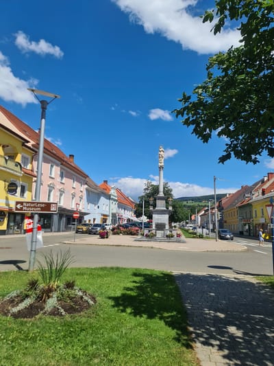 This is a street-level, full shot of a town square in Sankt Marein bei Neumarkt, Austria, on a clear, sunny day with scattered clouds. The perspective is from a sidewalk looking across a street intersection.

In the foreground to the left, there is a circular garden bed with green grass surrounding it, featuring some low-lying plants. To the right, a paved sidewalk runs parallel to the street, with dappled shade from trees overhead.

The midground is dominated by a cobblestone roundabout with a large monument in the center. The monument is a tall, stone column topped with a statue of a robed figure, possibly a religious icon. The base of the monument is surrounded by flower beds. Cars are visible driving on the road around the roundabout, and parked cars line the opposite side of the street.

The background features a row of colorful buildings lining the street, typical of Austrian town architecture. These buildings are of varying heights and colors, including yellow, pink, light blue, and green. Some have awnings and signs. Further in the distance, rolling green hills with scattered trees are visible under the bright blue sky. A street lamp is present in the left midground, and a traffic signal with a red light is visible on the right. A person in a blue dress is walking across a pedestrian crossing on the right side of the image.