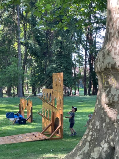 An outdoor scene at the Forest Park in Stupava, Slovakia features a temporary wooden castle-themed play structure on a grassy area. Two young boys are interacting with the structure. The boy on the left, wearing a grey t-shirt, blue shorts, a baseball cap, and sneakers, is looking upwards and appears to be engaged in an activity related to the structure. The second boy, partially obscured by a large tree trunk on the right, is wearing a green shirt and is also looking towards the structure. In the background, two individuals are walking, and several blue crates are visible near the play structure. The environment is characterized by lush green grass and numerous mature trees.
